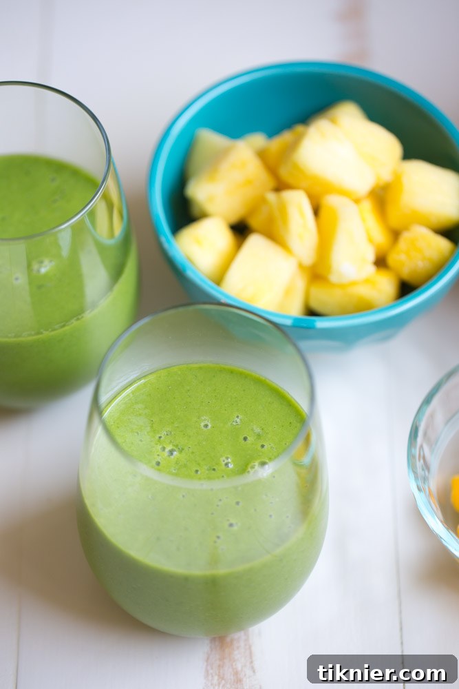 A vibrant green Tropical Kale Smoothie in a glass, garnished with fresh kale, mango, and pineapple chunks, set against a bright, clean background.