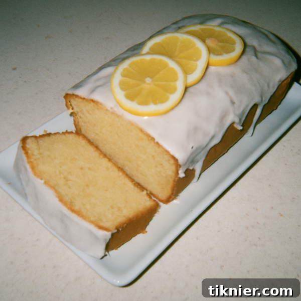 A perfectly glazed slice of homemade Starbucks Lemon Loaf on a plate, with a full loaf in the background, adorned with fresh lemon slices and zest.