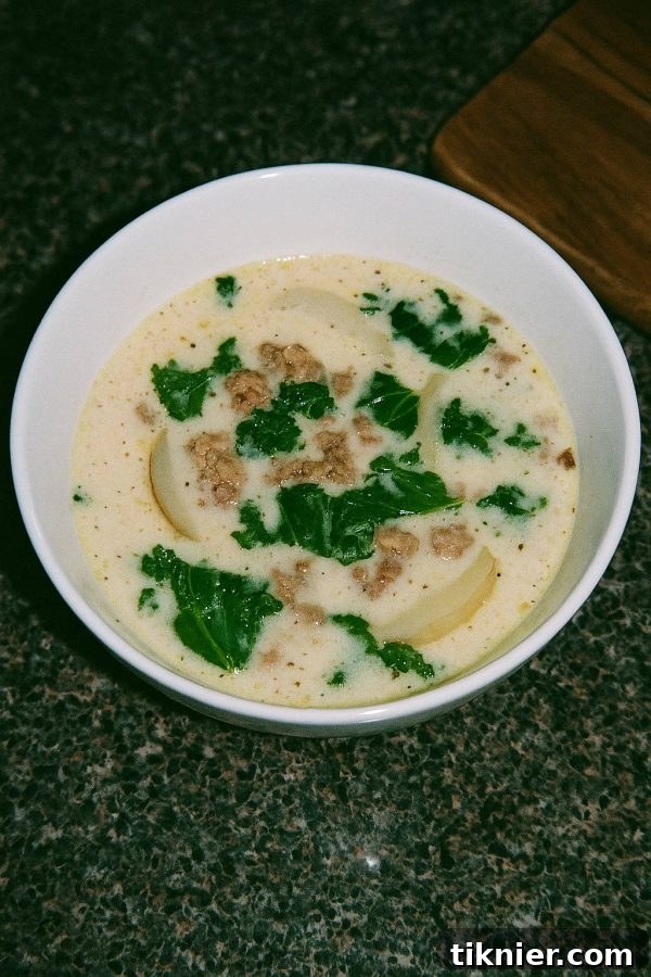 A steaming bowl of homemade Zuppa Toscana soup, garnished with fresh parsley and grated Parmesan cheese. The soup contains spicy Italian sausage, tender potatoes, and vibrant green kale in a creamy broth, with a rustic, cozy feel.
